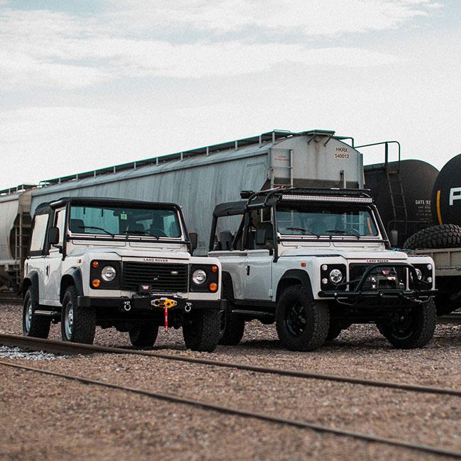Two Land Rover Defenders parked next to each other in a train yard